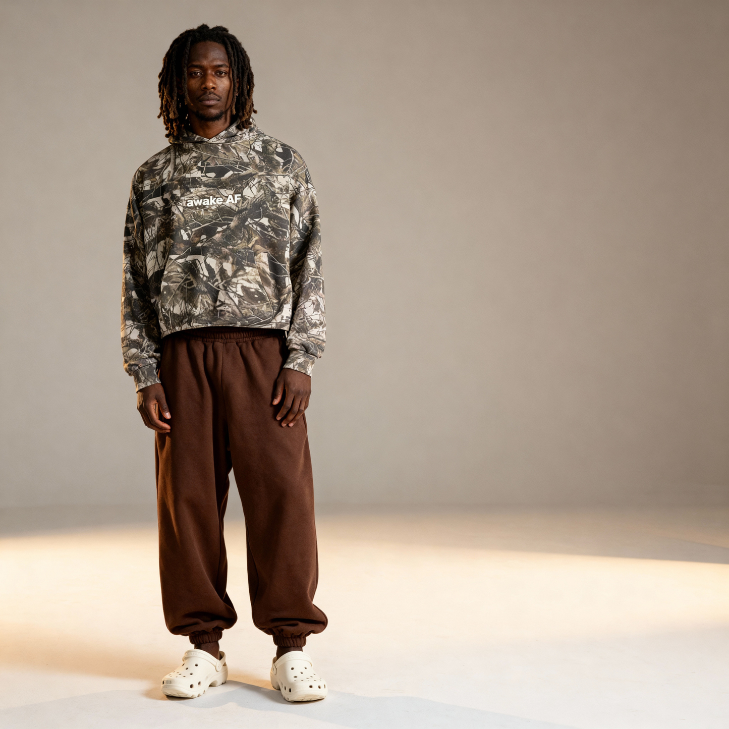 Person wearing a camouflage hoodie and brown pants on a neutral background