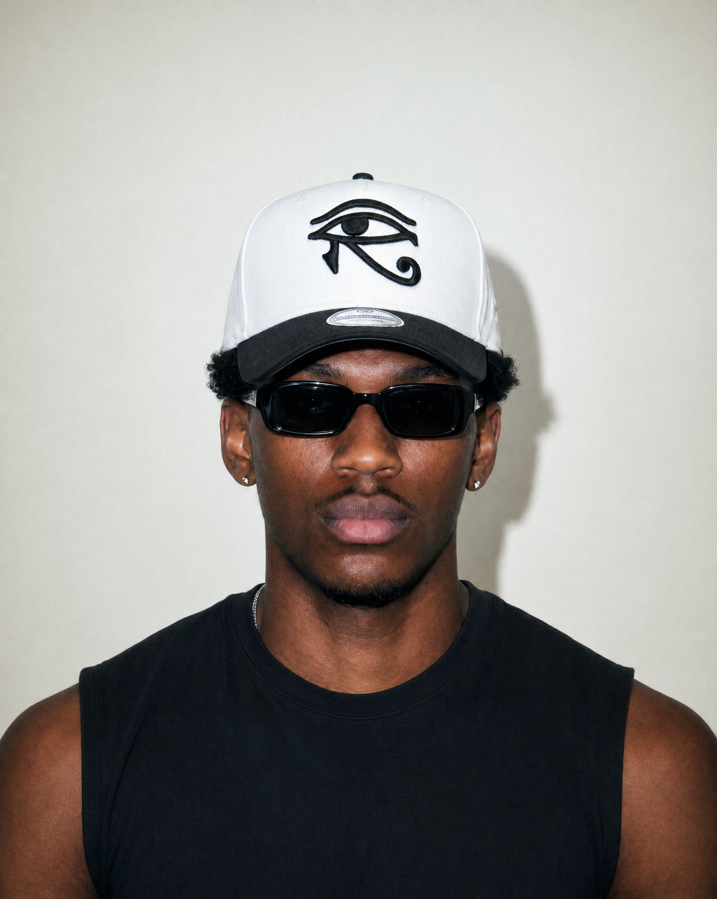 Man wearing a white cap with an eye symbol and sunglasses against a plain background