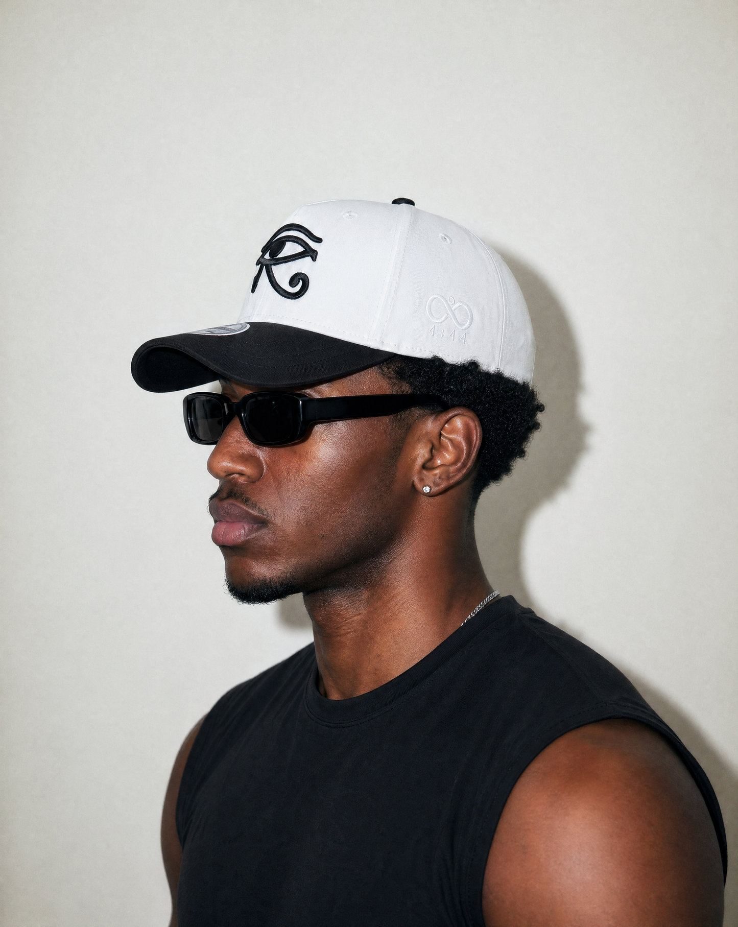 Man wearing a white and black cap with a logo and sunglasses, against a plain background