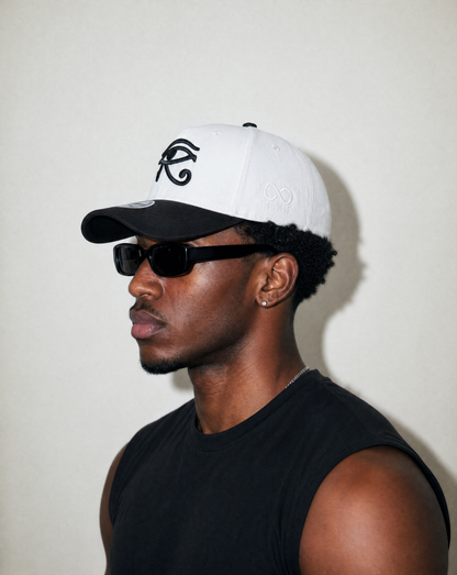 Man wearing a white and black cap with a logo and sunglasses, against a plain background